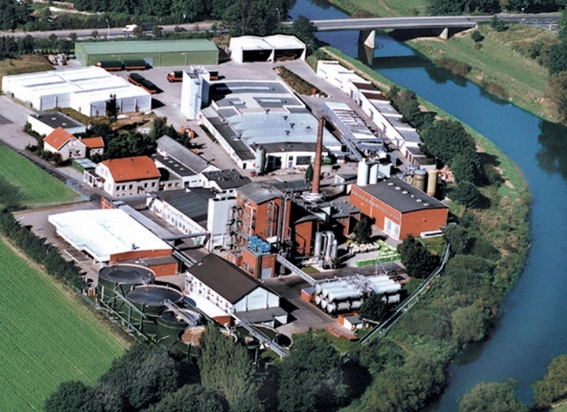 Luftaufnahme einer Industrie- und Kläranlagenanlage mit mehreren Gebäuden, Tanks und Schornstein entlang eines Flusses