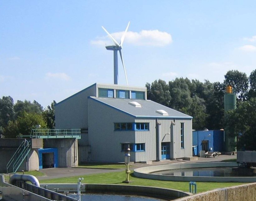 Gebäude und Rundbecken einer Kläranlage mit Windkraftanlage im Hintergrund im ländlichen Umfeld